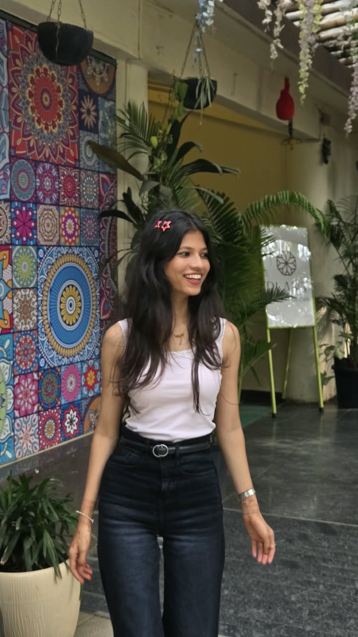 Bhakti laughing in a white top and dark jeans in front of a mandala-tile mural, a red star clip in her hair