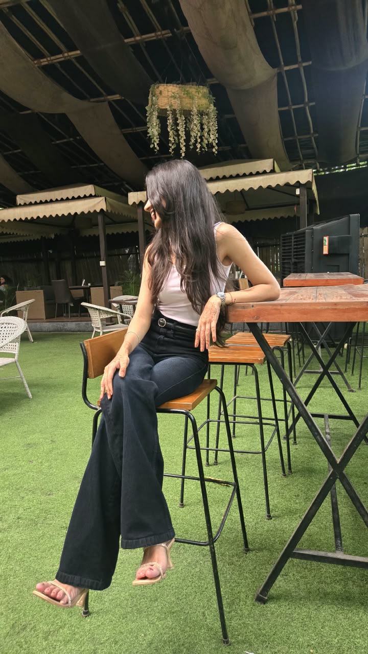 Bhakti in a white top and wide-leg jeans, seated on a wooden bar stool on artificial grass and looking away