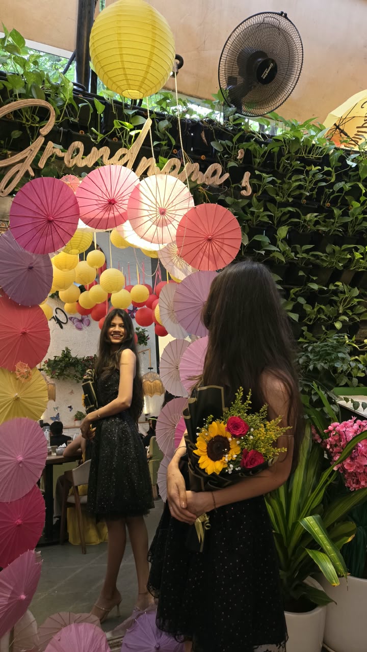 Bhakti in a black polka-dot dress with a sunflower bouquet, turned away beside pastel paper parasols and hanging plants at a café