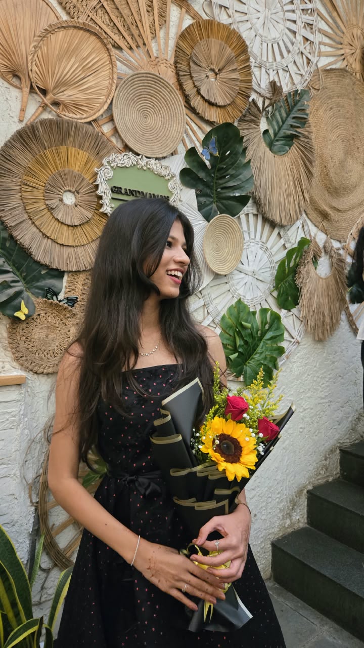 Bhakti laughing in a black polka-dot dress, holding a sunflower and rose bouquet in front of a wall of woven fans and leaves