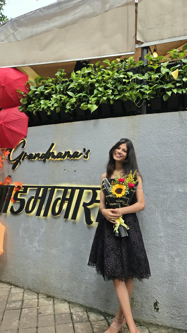 Bhakti smiling in a black polka-dot dress, holding a sunflower bouquet and leaning against a grey wall with a glowing café sign