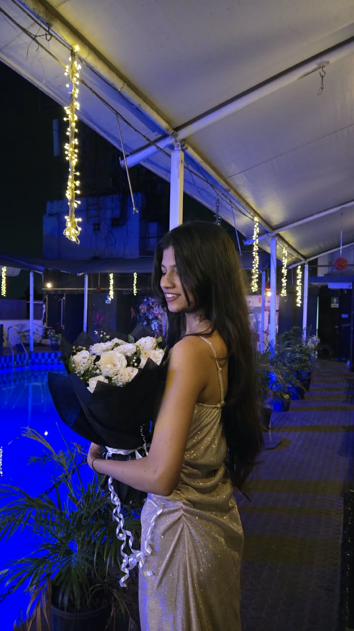 Bhakti in a gold shimmer dress glancing back over her shoulder with a bouquet of white roses, beside a night-lit rooftop pool