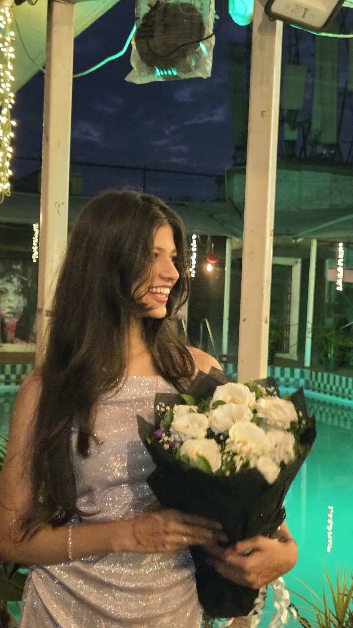 Close portrait of Bhakti laughing in a silver glitter dress, holding white roses by a glowing teal pool at night