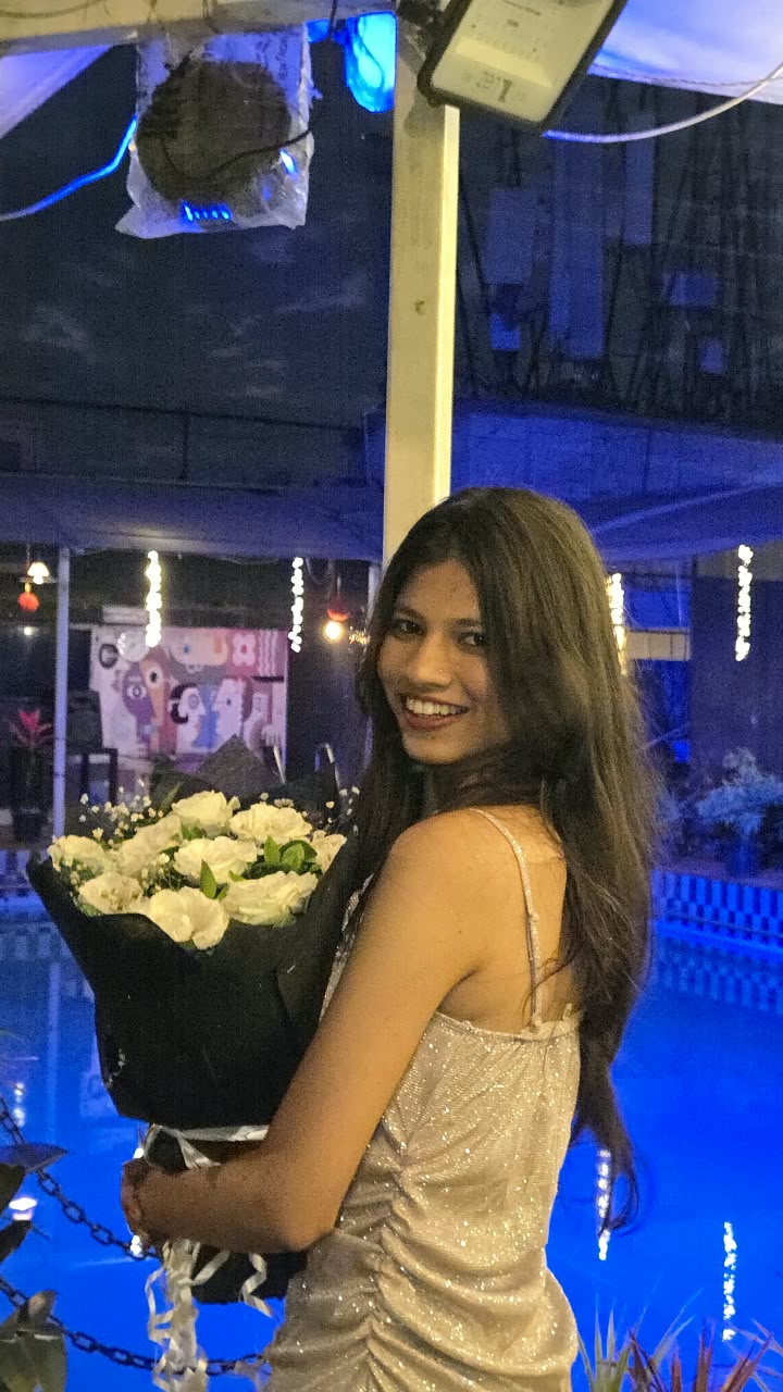 Bhakti smiling over her shoulder in a shimmery champagne dress with a bouquet of white roses, beside a blue-lit pool at night