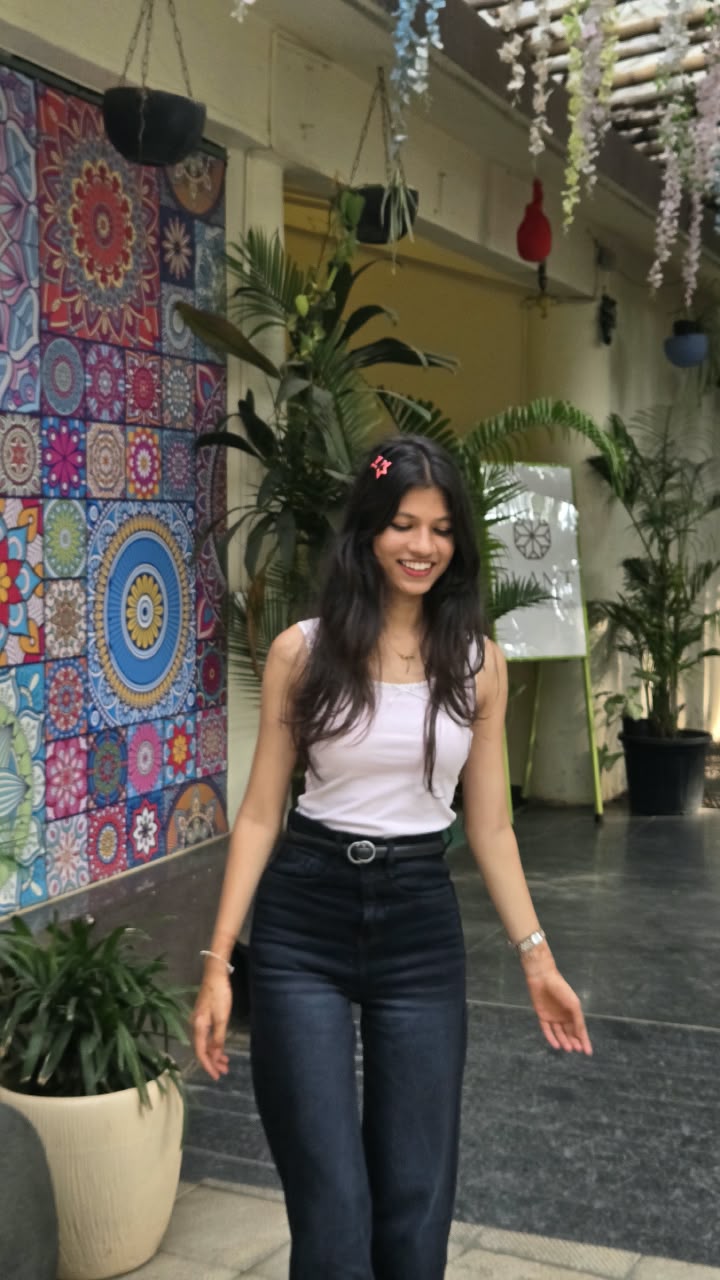 Bhakti in a white tank top and high-waisted dark jeans, walking past a colourful mandala-tile mural with a star clip in her hair