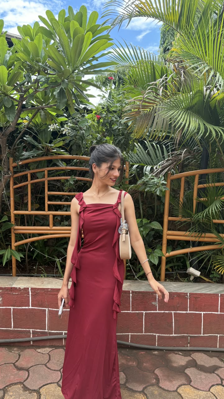 Bhakti in a burgundy ruffle-strap maxi dress with a small cream bag, standing in front of a bamboo trellis and tropical plants