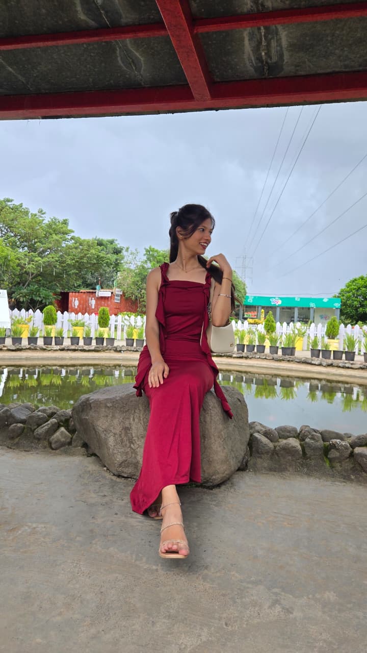 Bhakti in a burgundy maxi dress sitting on a boulder beside a small pond, glancing back over her shoulder