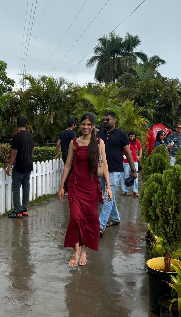 Bhakti in a burgundy maxi dress walking along a rain-wet garden path